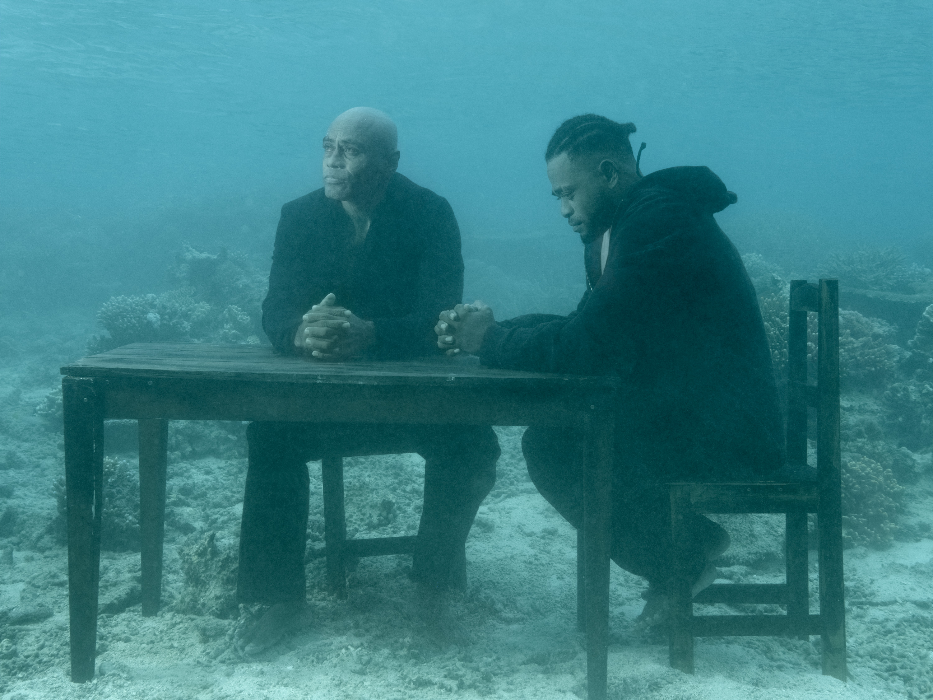 Two men sit at a wooden table on the ocean floor, surrounded by coral and clear blue water.