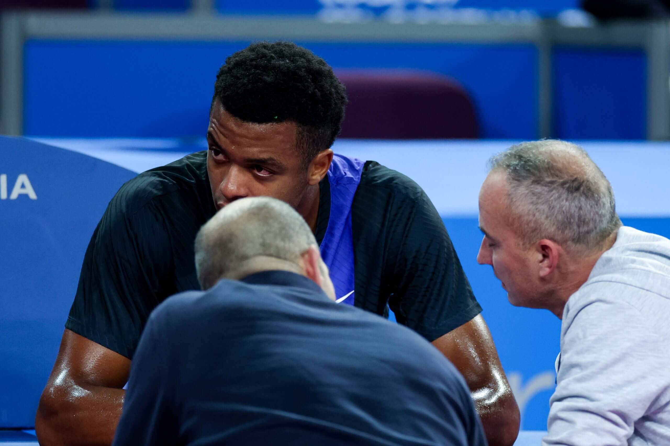 Giovanni Mpetshi Perricard sits at the side of a tennis court and looks on despondently while a tournament trainer and official talk to him.