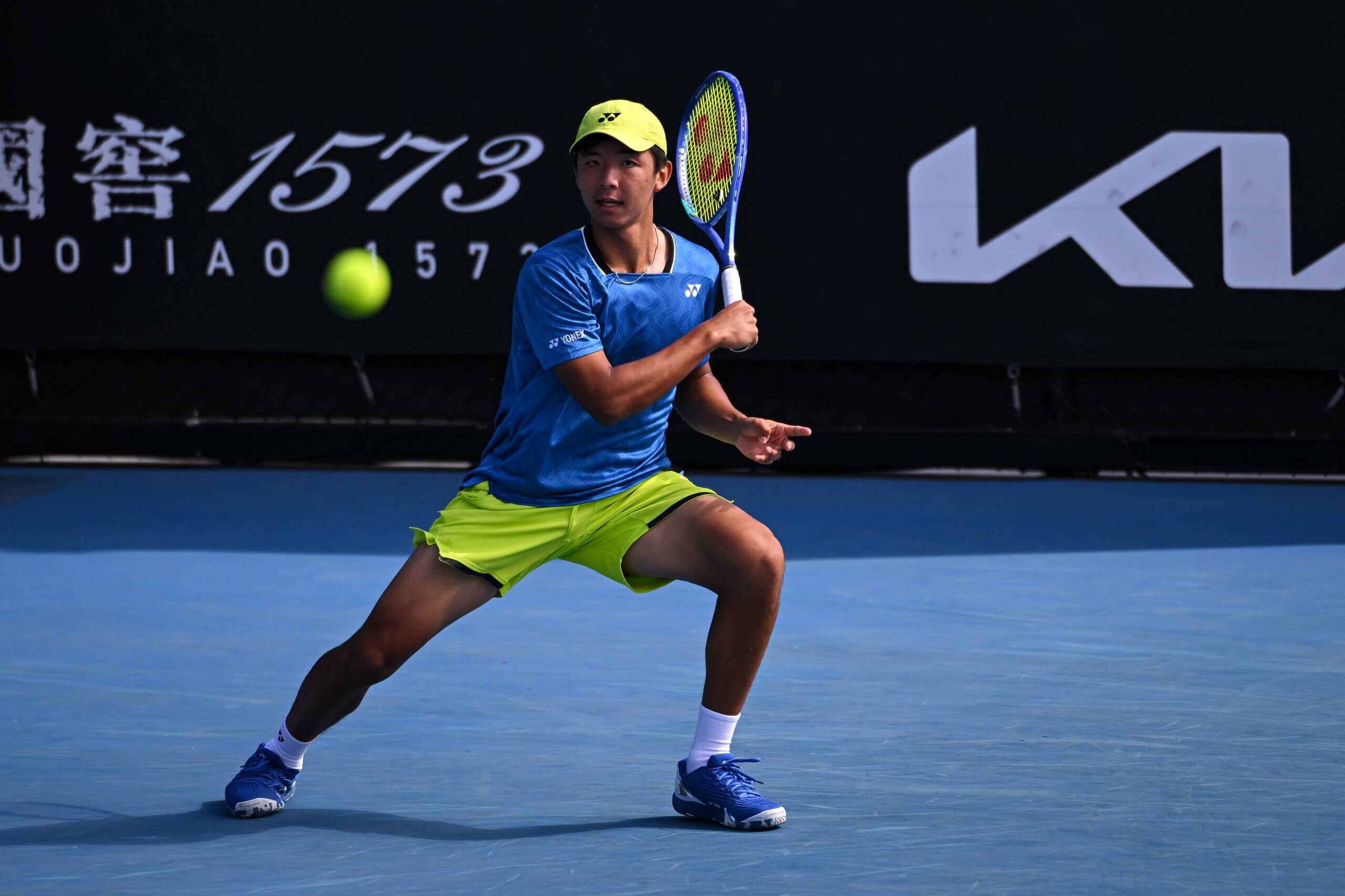 Zangar Nurlanuly hits a forehand on a blue tennis court.