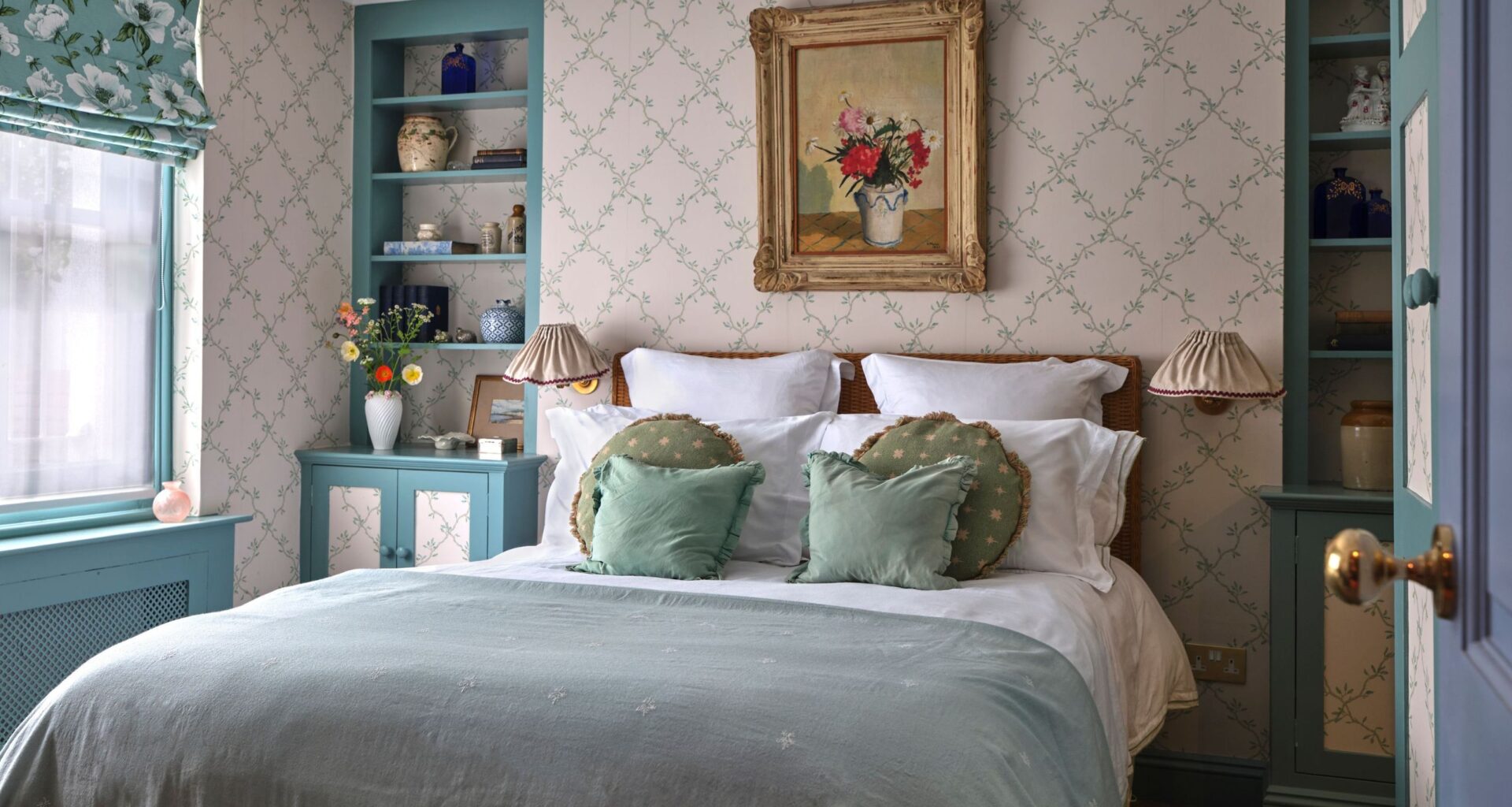 A bed with light blue linens and green decorative pillows set against a delicate floral trellis wallpaper. A teal built-in bookshelf sits to the right, filled with blue and white ceramics.