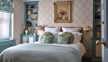 A bed with light blue linens and green decorative pillows set against a delicate floral trellis wallpaper. A teal built-in bookshelf sits to the right, filled with blue and white ceramics.