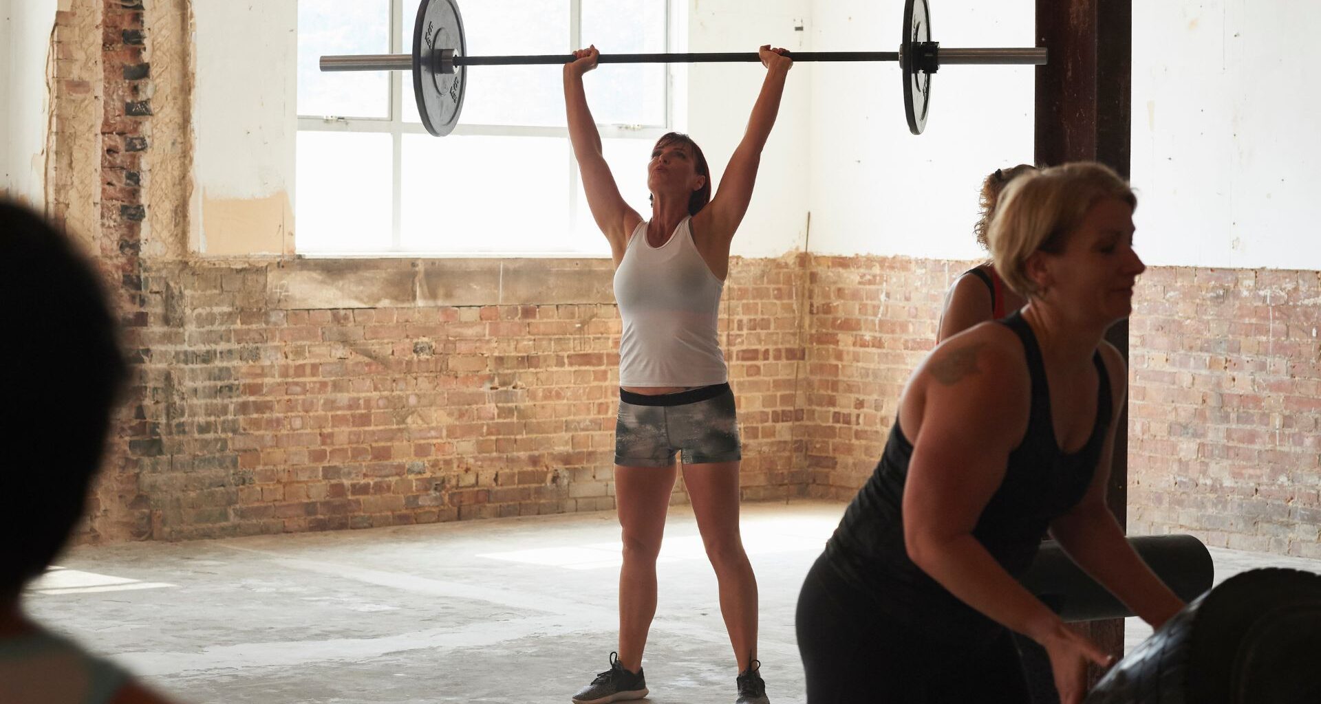 Woman lifting barbell above head with weight on either side wearing gym clothes and standing with friends in foreground
