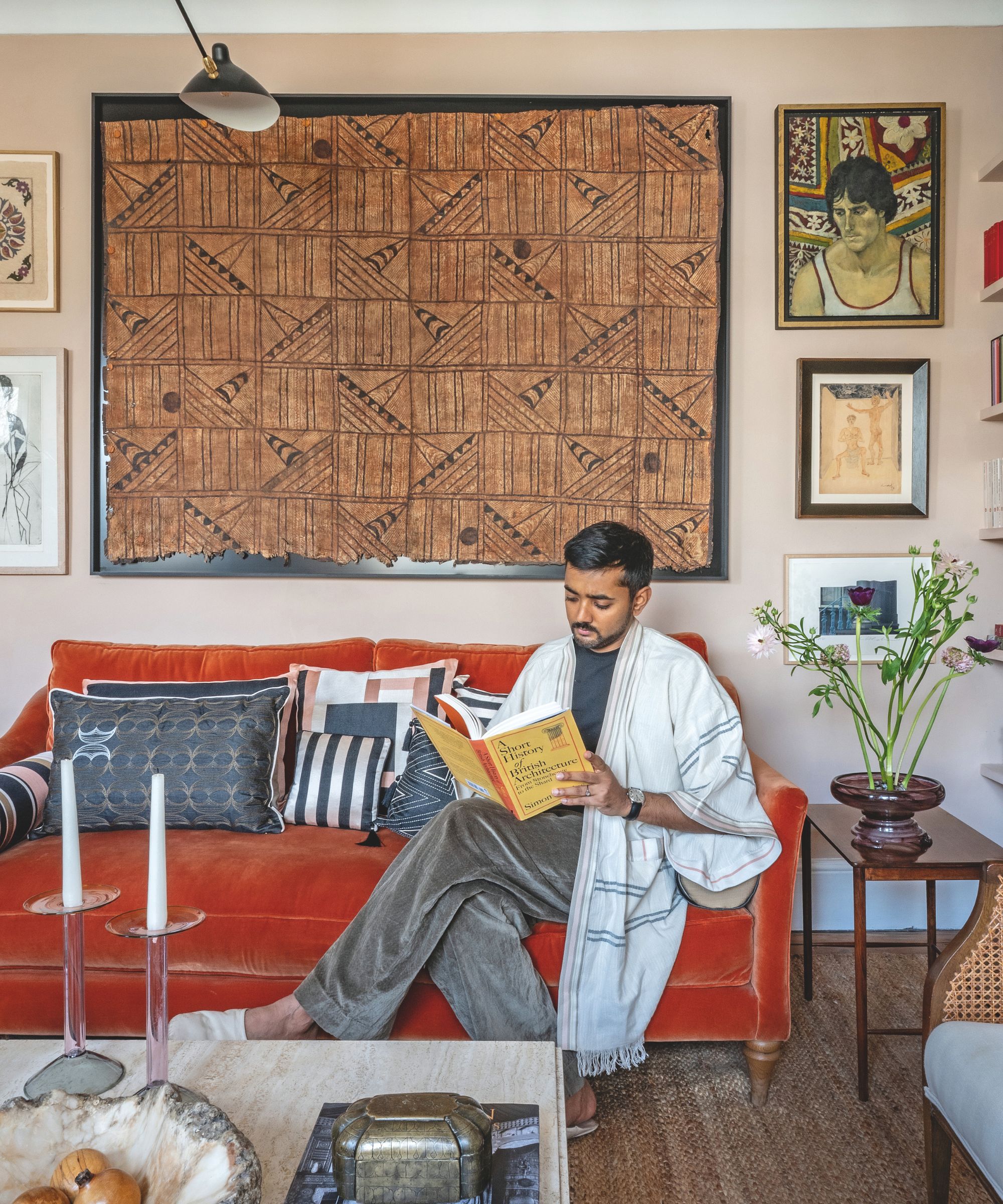 A man sits on an orange velvet sofa, wearing a white robe and reading a book titled "A Short History of British Architecture" beneath a large framed textile