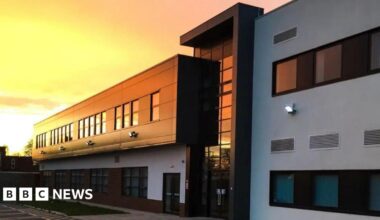 Image shows the outside of Melton Theatre which is a white and black modern looking building. The sun is setting over the building which is reflecting on the windows and giving it a warm yellow, orange glow.