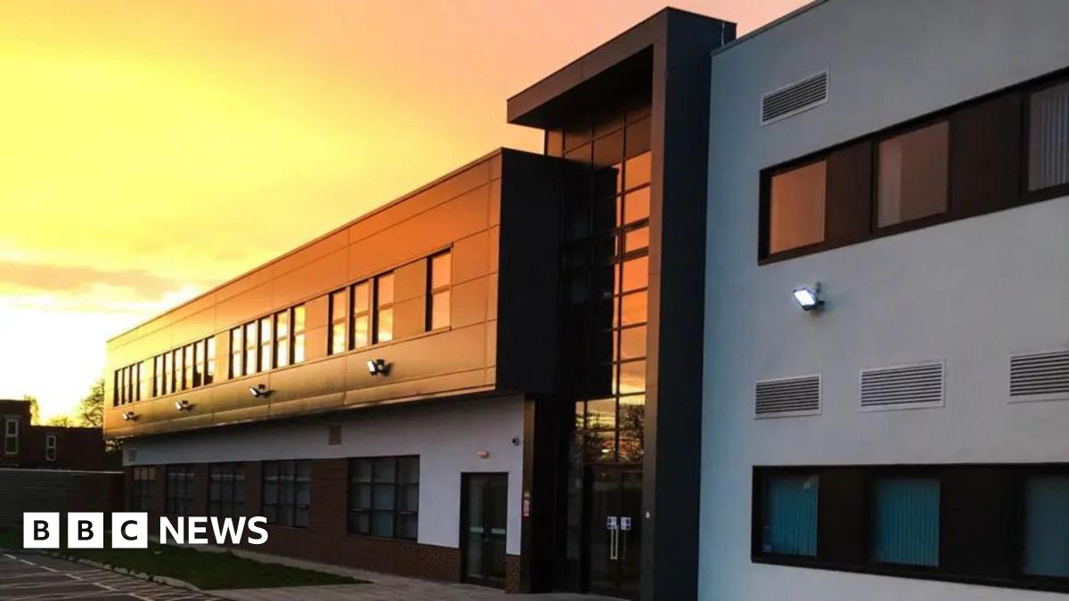 Image shows the outside of Melton Theatre which is a white and black modern looking building. The sun is setting over the building which is reflecting on the windows and giving it a warm yellow, orange glow.