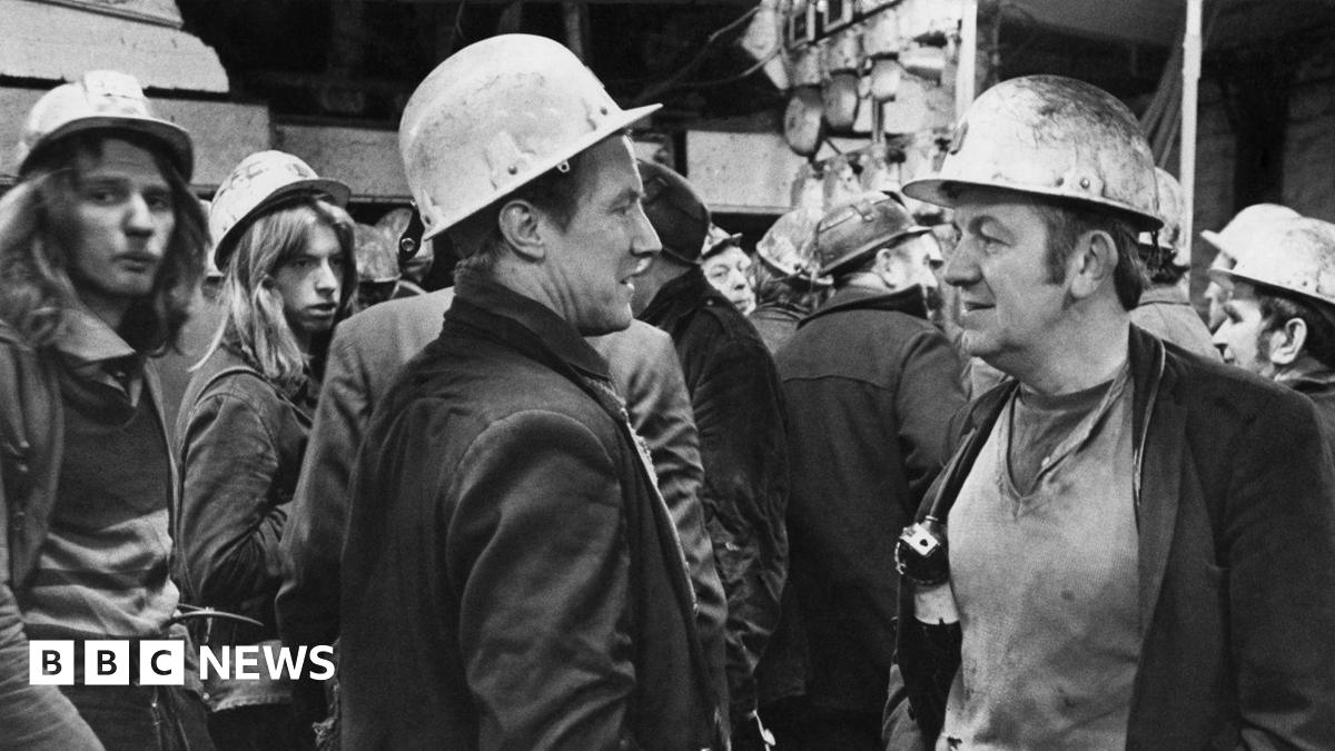 A black and white photograph of a group of men with hard hats on and in worn, dirty clothes. McGuinness stands on the right talking to another man. He has brown hair with large sideburns.