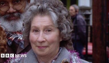 Three women and a man stood in a row smiling looking ahead. They are wearing jackets with coloured tinsel round their necks. They are stood outside and in the background is a tree with coloured lights on.