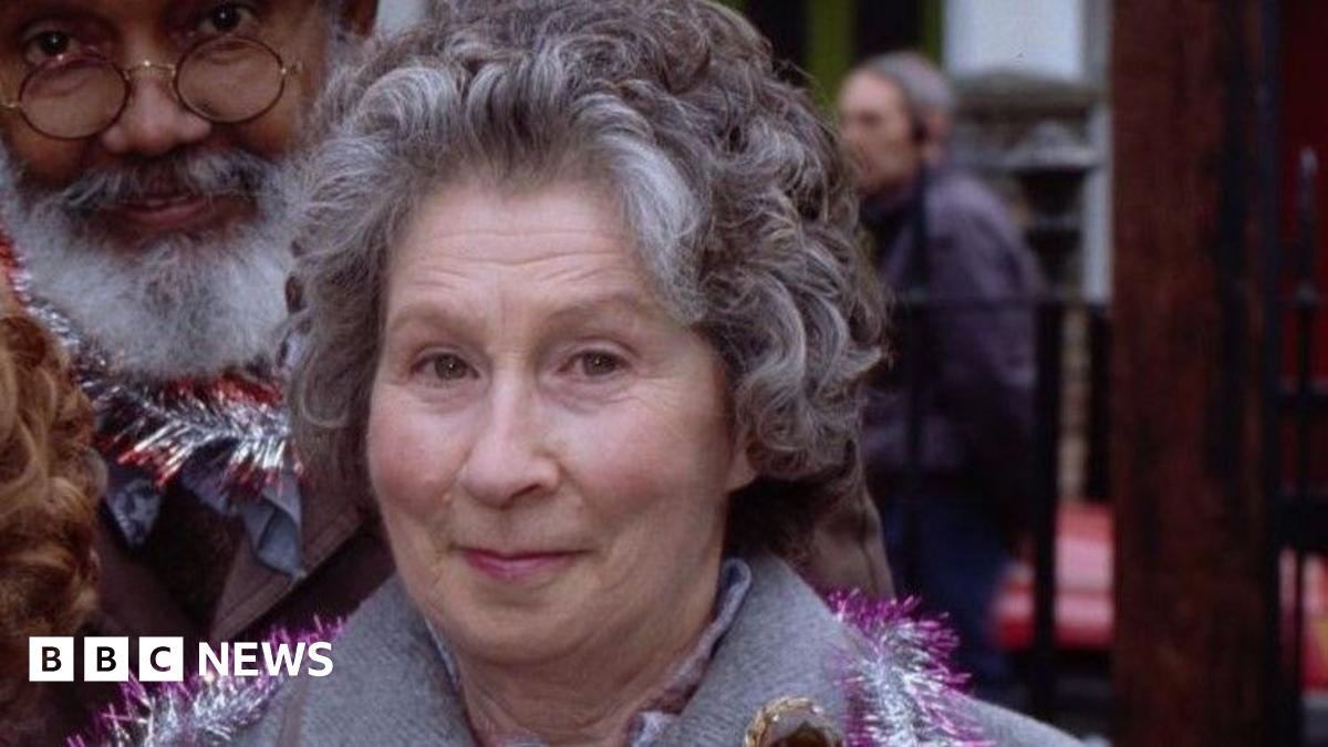Three women and a man stood in a row smiling looking ahead. They are wearing jackets with coloured tinsel round their necks. They are stood outside and in the background is a tree with coloured lights on.