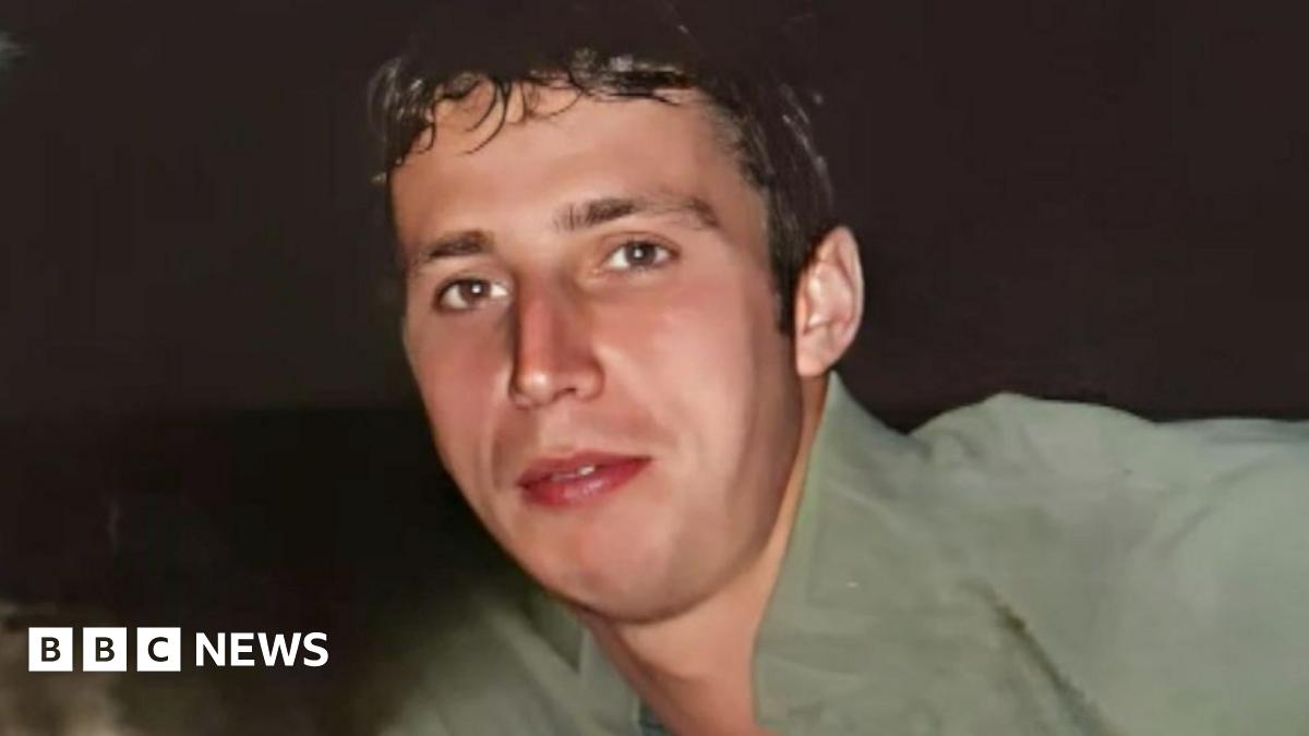 Aaron Armstrong pictured with a dog. The dog, which is white with black spots, is laying on Aaron's chest. Aaron is pulling a kissing face at the dog, and is wearing a white t-shirt.