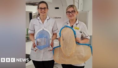 Two nurses are standing next to each other. On the left the woman has brown hair and glasses, and is wearing a white uniform. On the right the woman has blonde hair and glasses, with white uniform and a yellow badge. On the left the woman is holding a small mask that goes over someone's face - it has blue mesh that covers much of the patient's face but with a large hold for their nose and mouth. On the right, the mask is mesh and would cover the entirety of someone's head and shoulders