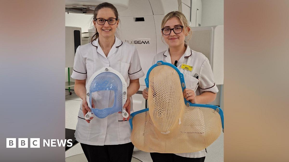 Two nurses are standing next to each other. On the left the woman has brown hair and glasses, and is wearing a white uniform. On the right the woman has blonde hair and glasses, with white uniform and a yellow badge. On the left the woman is holding a small mask that goes over someone's face - it has blue mesh that covers much of the patient's face but with a large hold for their nose and mouth. On the right, the mask is mesh and would cover the entirety of someone's head and shoulders