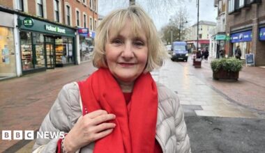 A blonde woman wearing a light grey coat and a red scarf. She is standing in the middle of a high street.