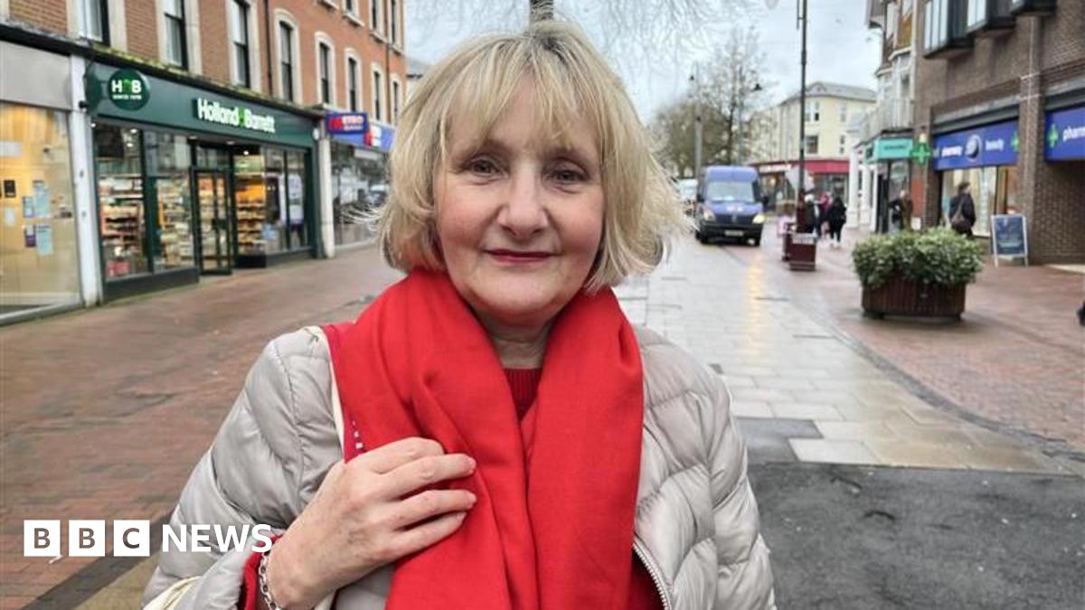 A blonde woman wearing a light grey coat and a red scarf. She is standing in the middle of a high street.