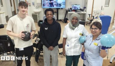 Two men and two women inside a hospital stroke recovery ward. There are wheelchairs behind them, along with physiotherapy exercise balls, computers and a large TV screen.