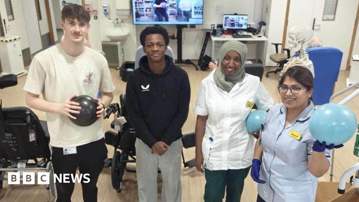 Two men and two women inside a hospital stroke recovery ward. There are wheelchairs behind them, along with physiotherapy exercise balls, computers and a large TV screen.