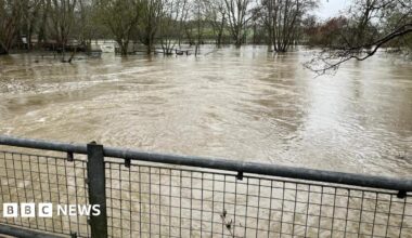 A river which has risen significantly and is a brown colour. There is a metal fence in the way.
