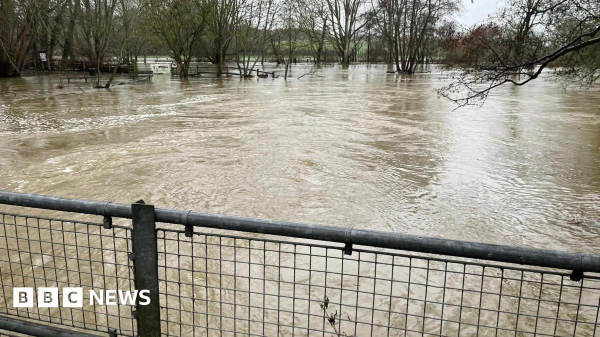 A river which has risen significantly and is a brown colour. There is a metal fence in the way.
