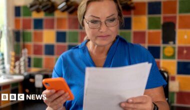 Woman in a kitchen holds a smartphone in one hand and a bill in the other, with colourful tiles on the wall behind her.