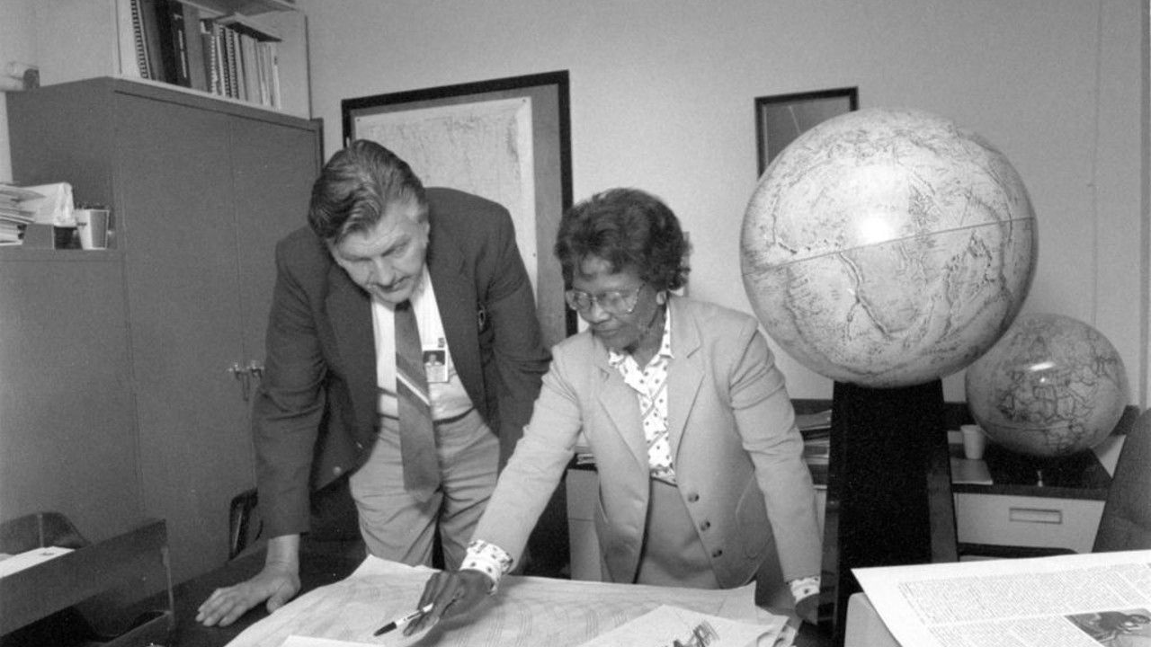 a woman in a suit points at a map on a table next to a globe