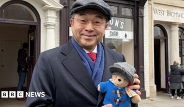 Hiroshi Suzuki has a grey flat cap on and a navy blue coat, with a blue scarf and red tie. He wears glasses and is holding a small Paddington Bear toy which also has a grey flat cap on, and a blue coat.