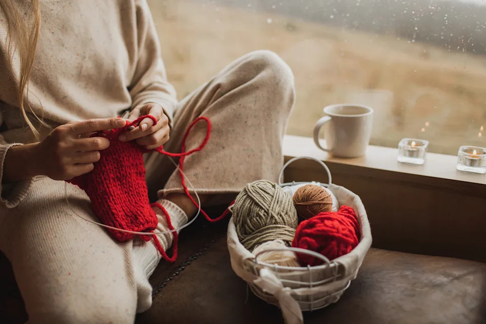 Person knitting with yarn by a window, cozy ambiance, cup, and candles nearby. Image illustrates relaxation and creative work