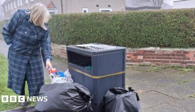 Joan McTigue is wearing a long blue coat and looking at a black bin surrounded with black bin bags full of rubbish.
