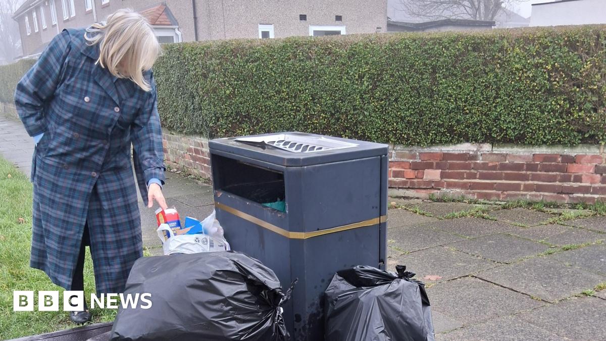 Joan McTigue is wearing a long blue coat and looking at a black bin surrounded with black bin bags full of rubbish.