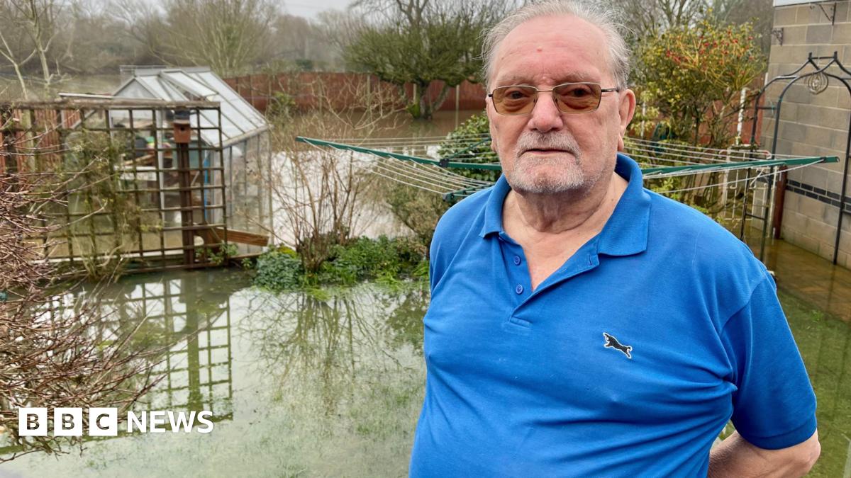 A man in a blue polo shirt and dark glasses is standing in a flooded domestic garden. Bushes, plants, a garage, greenhouse and washing pole is partially submerged. There is a wood panelled fence around the garden. Trees stand behind the fence in a field which is also flooded.