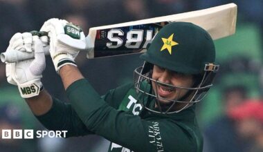 Pakistan's Saim Ayub plays a shot during the third T20 international cricket match between Pakistan and Australia. He is wearing a green helmet that has a wire face guard. His shirt is also dark green while his gloves are white. His bat is raised up around his head.