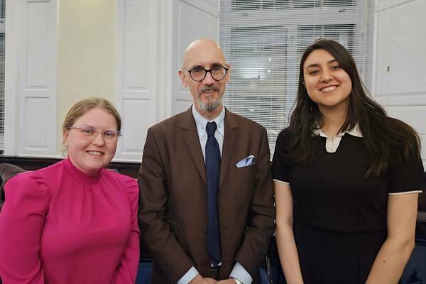 Three smiling people. A blonde woman with glasses in a fuchsia ruffled blouse, a bald man with a grey beard and glasses in a brown suit and blue tie, and a dark-haired woman in a black top with white collar, stand indoors.