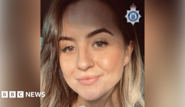 A woman with long, light brown hair smiles as she takes a selfie.