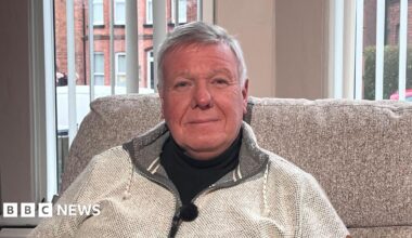 A man with short grey hair is pictured sitting in a living room in front of a large window. He is dressed in a black t-shirt with a light grey zip up jacket on top. He's sitting on a grey sofa. Behind him you can see red brick houses, a red car, and a white van outside the window.