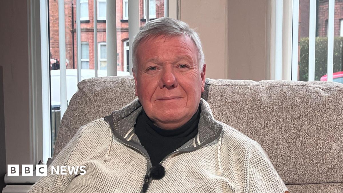 A man with short grey hair is pictured sitting in a living room in front of a large window. He is dressed in a black t-shirt with a light grey zip up jacket on top. He's sitting on a grey sofa. Behind him you can see red brick houses, a red car, and a white van outside the window.