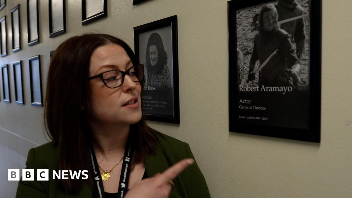 A woman stands in a school corridor lined with framed photographs displayed in neat rows along the wall. She has dark-brown, shoulder-length hair and is wearing glasses, a gold necklace and a green jacket with a lanyard. She is pointing towards one of the framed portraits beside them. It shows an actor with long hair holding a sword in the TV show Game of Thrones.