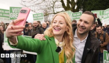 Hannah Spencer and Zack Polanski pose together as she takes a selfie with her pink mobile phone. A crowd of people stand behind in a park holding Vote Green placards