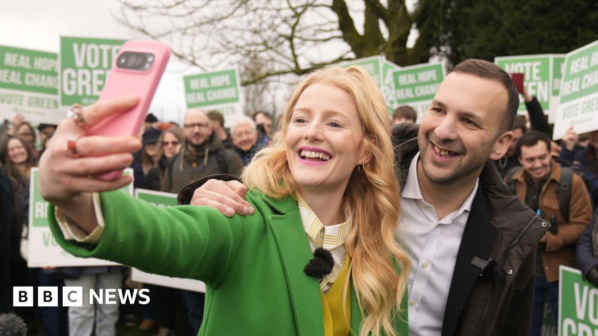 Hannah Spencer and Zack Polanski pose together as she takes a selfie with her pink mobile phone. A crowd of people stand behind in a park holding Vote Green placards