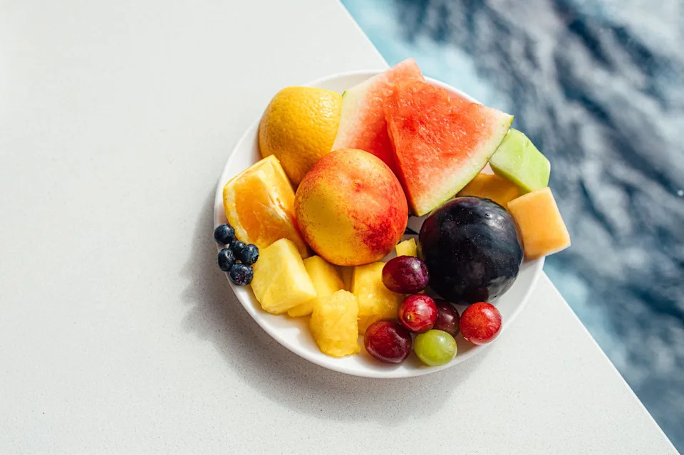 a colorful platter of assorted fruits including watermelon, peach, and grapes. set on a white surface, the fruits contrast beautifully with the blurred blue background.