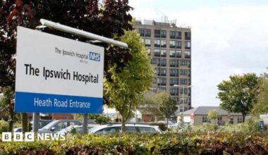 The entrance to Ipswich Hospital. A large white sign details the hospital and has a blue and white NHS sign on it. Cars can be seen behind the sign parked in a car park. A multi-storey building and other smaller buildings are in the background.