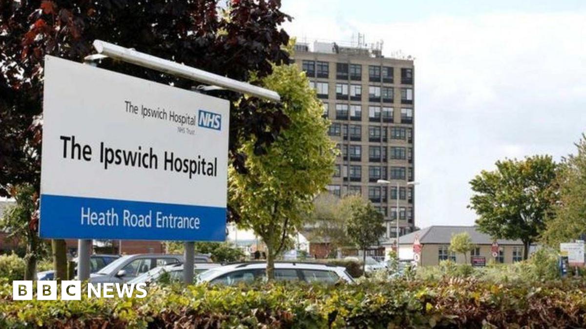 The entrance to Ipswich Hospital. A large white sign details the hospital and has a blue and white NHS sign on it. Cars can be seen behind the sign parked in a car park. A multi-storey building and other smaller buildings are in the background.