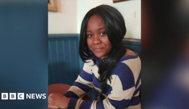 Chanel Thompson sits at a table in a café, wearing a blue and white striped top, looking at the camera.