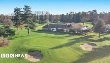 One of the putting greens and two bunkers on the golf course, which is dotted with fir trees. In the middle of the photo is a single storey building surrounded by a low hedge, with a car park next to it.