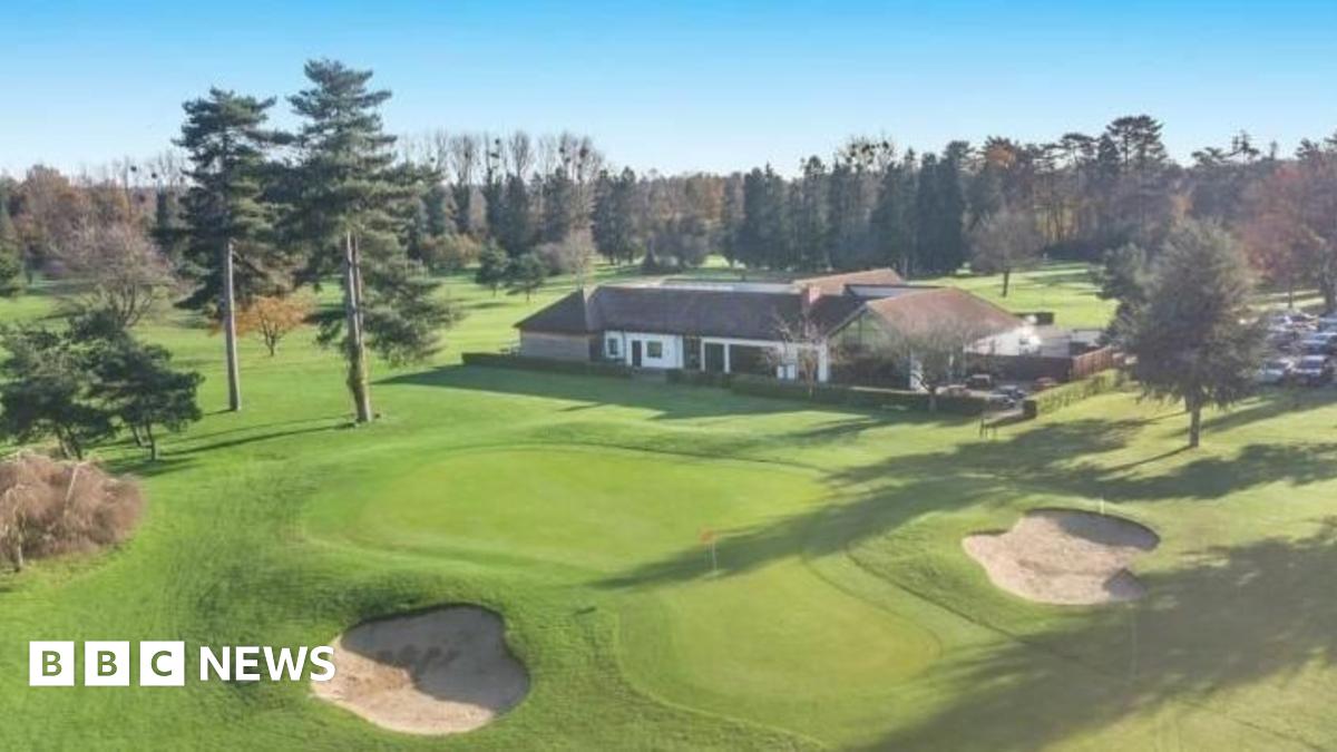 One of the putting greens and two bunkers on the golf course, which is dotted with fir trees. In the middle of the photo is a single storey building surrounded by a low hedge, with a car park next to it.