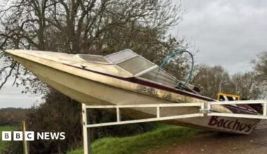 A cream coloured boat, shaped like a retro speedboat, leans against white barriers with its nose in the air, on a country lane leading into a field.