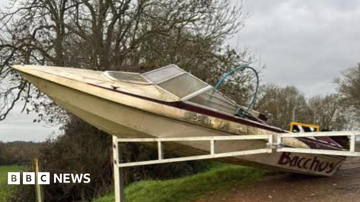 A cream coloured boat, shaped like a retro speedboat, leans against white barriers with its nose in the air, on a country lane leading into a field.