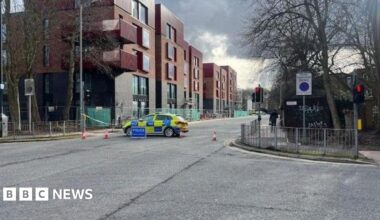 A shot of a street crossing, there is a police care across the road, as well as cones and police tape blocking the left exit off the road. To the far left there are buildings, with red brick at the top and black bricks at the bottom.