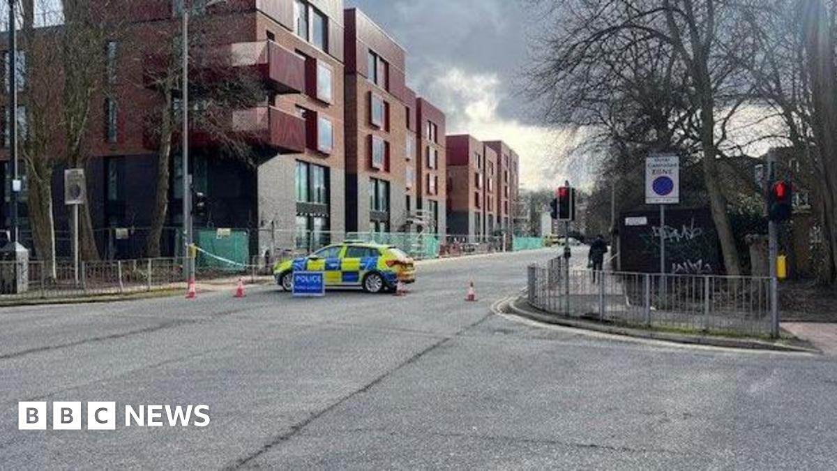 A shot of a street crossing, there is a police care across the road, as well as cones and police tape blocking the left exit off the road. To the far left there are buildings, with red brick at the top and black bricks at the bottom.