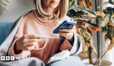 Sick woman at home wrapped in blanket measuring temperature and talking on smartphone for medical consultation.