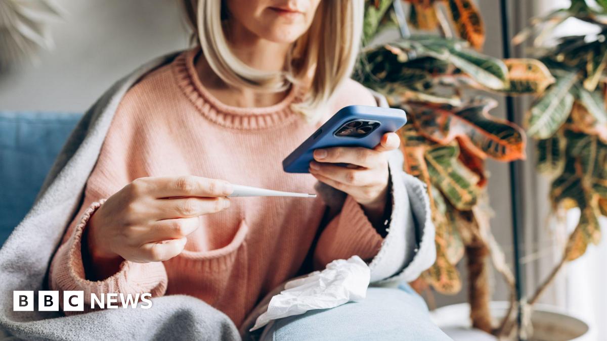 Sick woman at home wrapped in blanket measuring temperature and talking on smartphone for medical consultation.