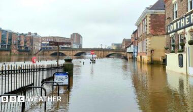 picture of the River Ouse in York flooded over the banks onto the pavement and road with floodwater rising up the side of the buildings.