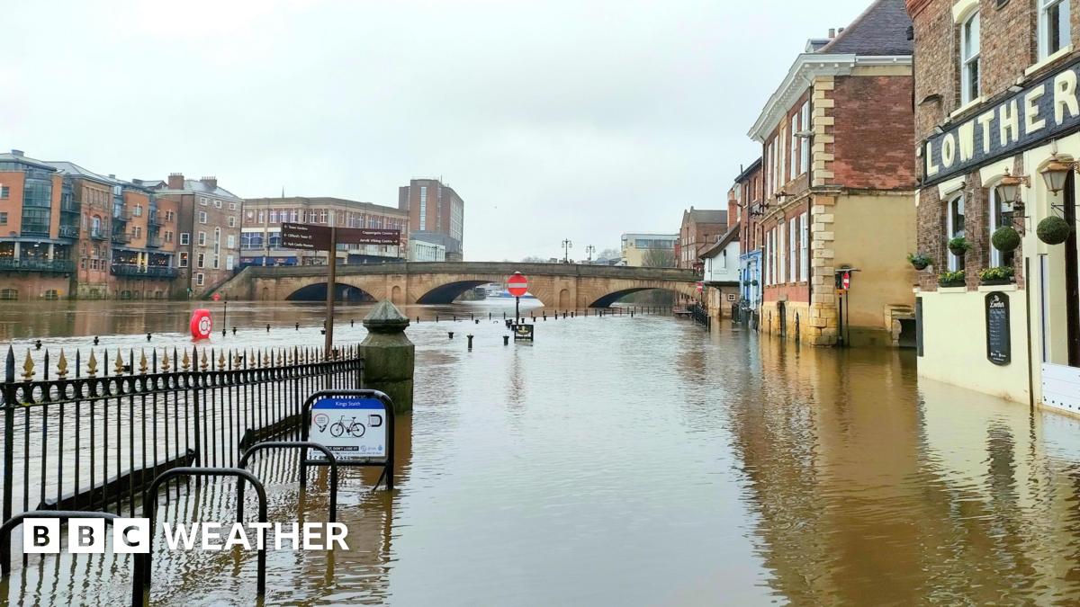 picture of the River Ouse in York flooded over the banks onto the pavement and road with floodwater rising up the side of the buildings.
